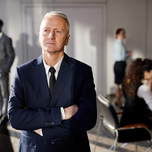 Waist up portrait of pensive senior man standing with arms crossed in conference room, copy space