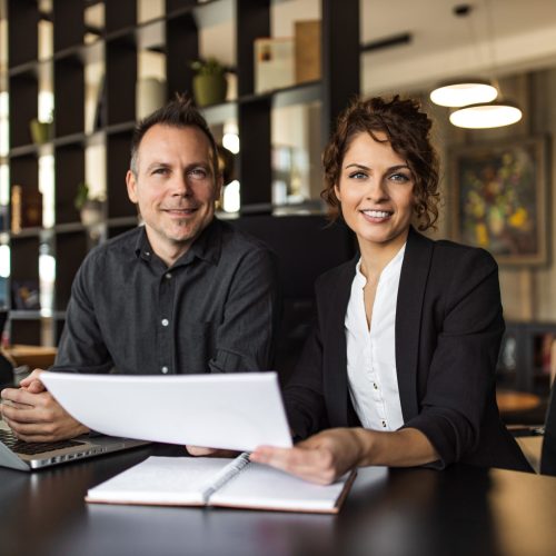 Portrait of a two cheerful HR managers at work. Sitting at desk in the office, holding paper document and looking at camera.