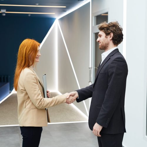 Portrait of successful red haired businesswoman shaking hands with smiling male colleague while standing in futuristic office interior, copy space